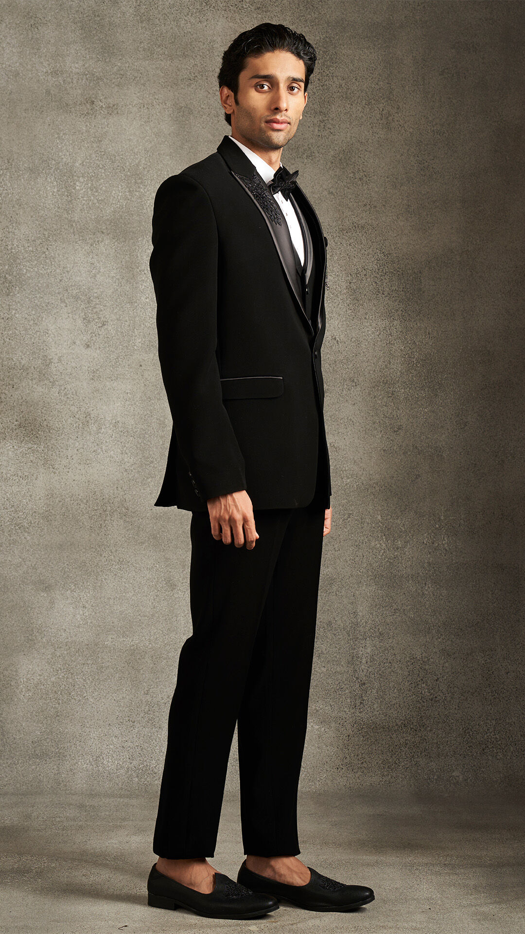 Man in a black tuxedo jacket with satin lapels, a waistcoat, and black loafers, posing formally.